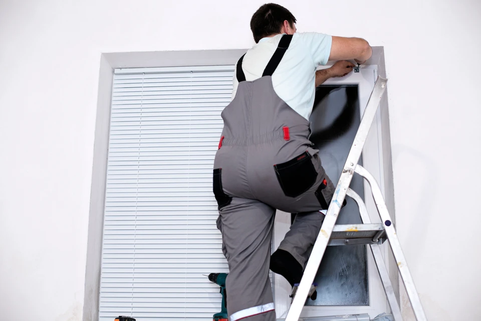 Homme en salopette grise et t-shirt blanc installant ou réparant un volet roulant intérieur depuis un escabeau