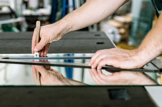 Atelier de menuiserie avec artisan manipulant une r&egrave;gle gradu&eacute;e sur panneau de verre r&eacute;fl&eacute;chissant