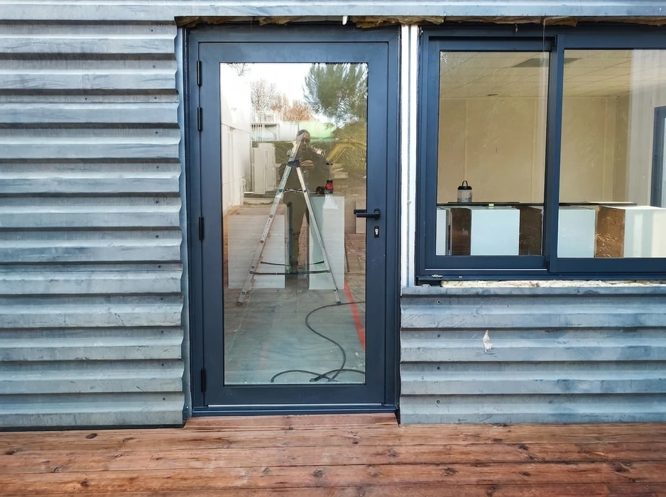 Technicien photographié en reflet dans une porte vitrée pleine hauteur en aluminium anthracite (RAL 7016), posée sur bardage métallique ondulé galvanisé