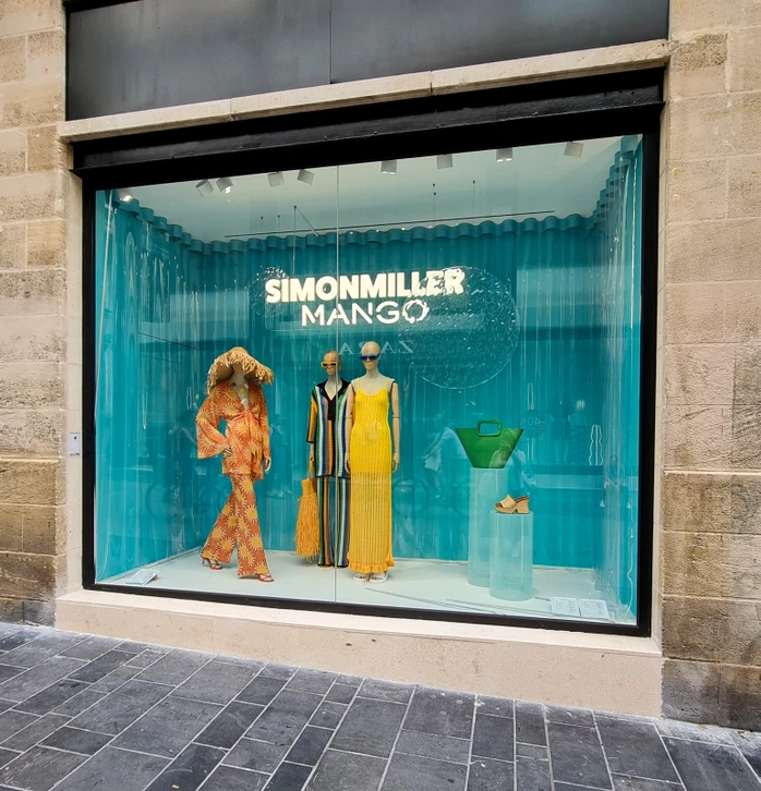 Photographie d'une équipe d'ouvriers spécialisés en miroiterie travaillant sur la façade vitrée d'un magasin ou d'un immeuble commercial. La scène montre des artisans en tenue professionnelle (casques, gilets de sécurité) manipulant des panneaux de verre ou des profilés métalliques, avec du matériel de chantier adapté. La façade en arrière-plan doit présenter une belle vitrine commerciale moderne avec vitrages épurés. La photo doit transmettre professionnalisme, expertise et envergure d'intervention sur des chantiers importants.