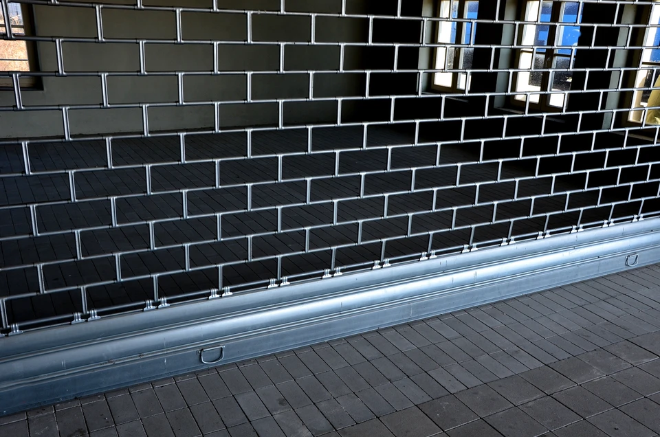 Devanture d'un local commercial ou magasin avec un rideau métallique de fermeture de couleur grise ou argent entièrement abaissé, bien posé et régulier, protégeant la vitrine. L'image montrera la façade du bâtiment en plein jour avec ciel clair, mettant l'accent sur l'installation soignée et la robustesse du système de fermeture. Pas de personne, pas de texte visible.
